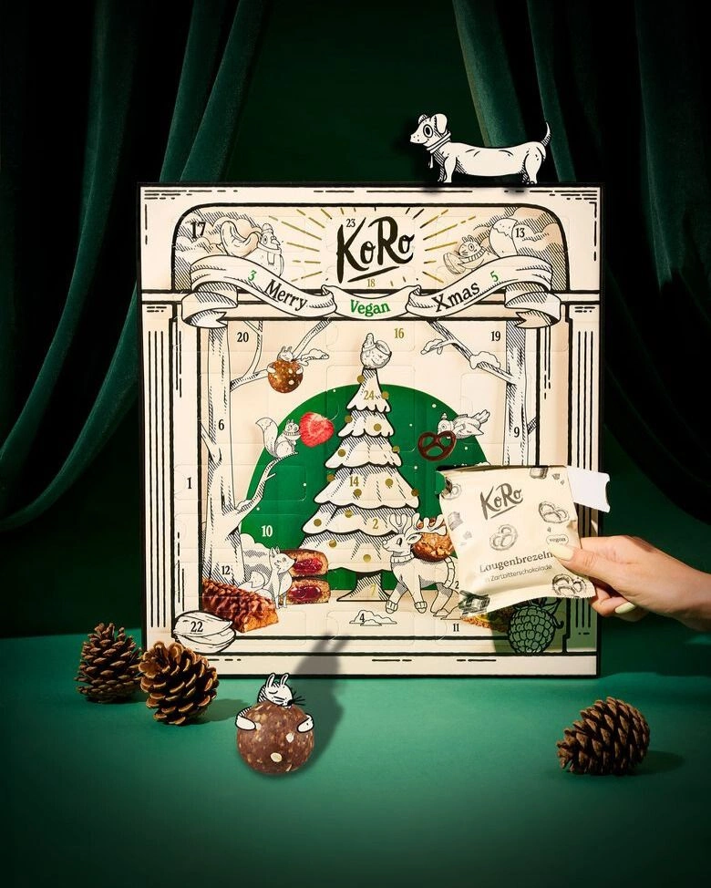 Vor einem grünen Hintergrund samt grünem Vorhang steht ein beigefarbener Adventskalender, auf dem ein gezeichneter Tannenbaum und Tiere zu sehen sind. Vor dem Kalender hält eine weibliche Hand ein Päckchen mit Laugenbrezeln in der Hand. Neben dem Kalender liegen Zapfen und ein Energy Ball.