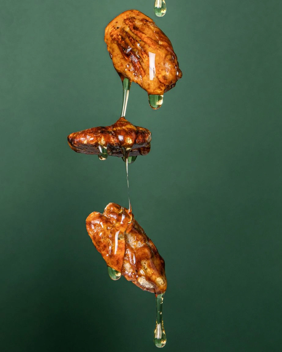 Drei goldbraune Pekannuskerne schweben in der Luft vor einem tiefgrünen Hintergrund. Sie sind mit klarem, glänzendem Karamell überzogen, das von der obersten Nuss über die mittlere bis zur untersten tropft. 