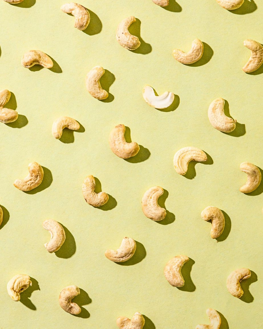 Einzelne Cashewkerne liegen in Reihen angeordnet auf einem hellgrünen Hintergrund. Die Cashewkerne sind ungeröstet und haben eine helle, cremeweiße Farbe. Sie werfen durch eine starke Lichtquelle von oben rechts Schatten.