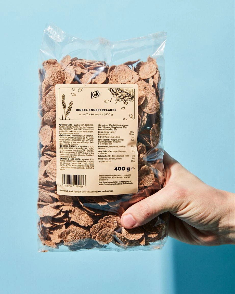 Eine Hand hält eine transparente Plastikverpackung von KoRo Dinkel Knusperflakes. Das Etikett der Verpackung zeigt die Nährwerttabelle, Zutatenliste und den Claim "Ohne Zuckerzusatz". Der Hintergrund ist hellblau.
