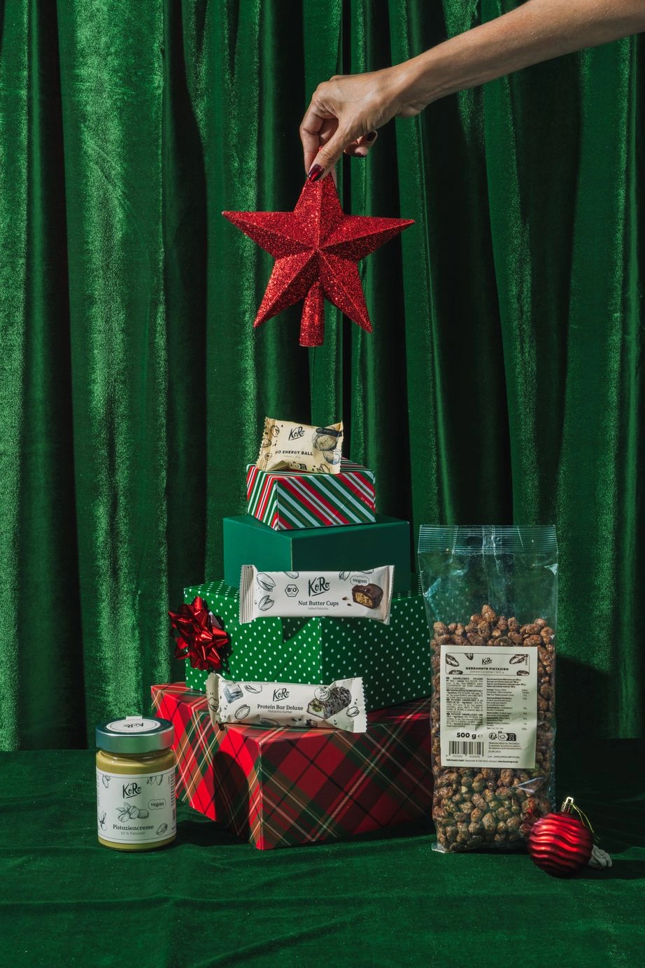 Eine festliche Weihnachtsinszenierung vor grünem Samthintergrund. Geschenkboxen in verschiedenen Grüntönen und Rot, gestapelt in Form eines Tannenbaums mit einem roten Stern als Spitze, umgeben von veganen Koro-Produkten wie Nussmus, Proteinriegeln, Nut Butter Cups und gerösteten Kichererbsen.