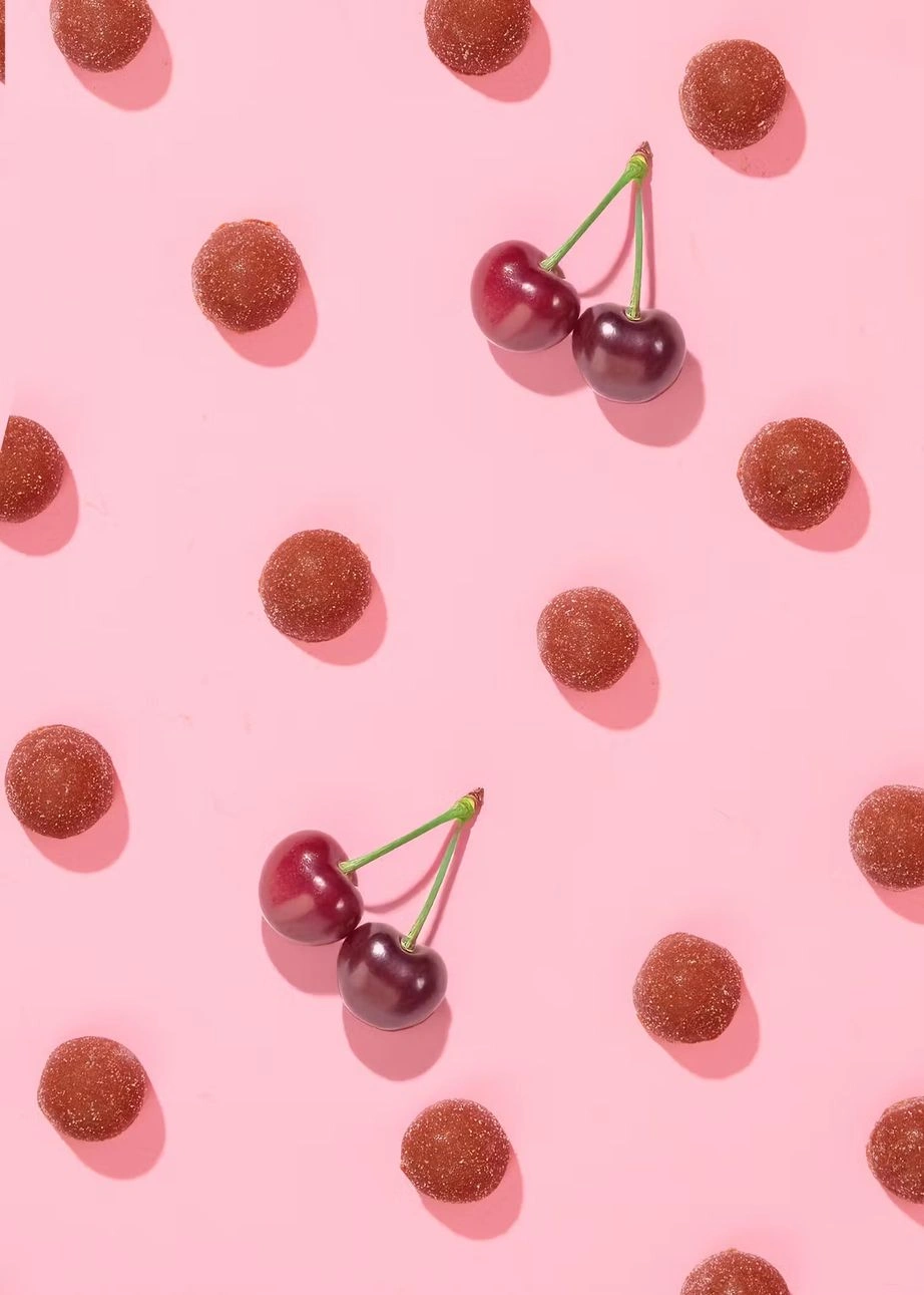 Ansicht der runden Frucht-Bites zusammen mit frischen Kirschen als dekoratives Pattern auf rosa Grund.