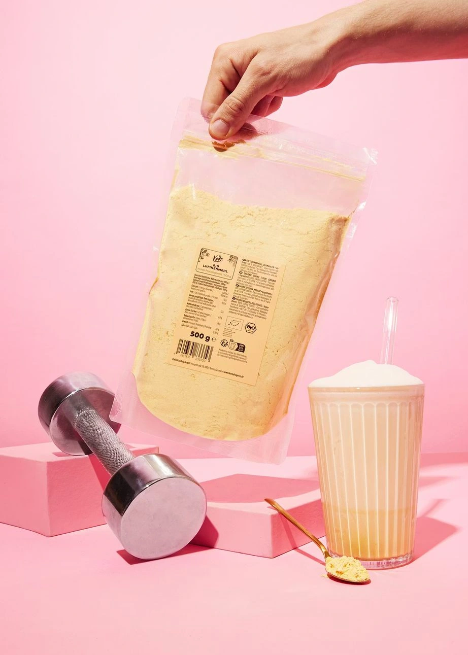 Eine Hand hält eine Packung KoRo Bio Lupinenmehl neben einer Hantel und einem Protein-Shake auf rosa Hintergrund.