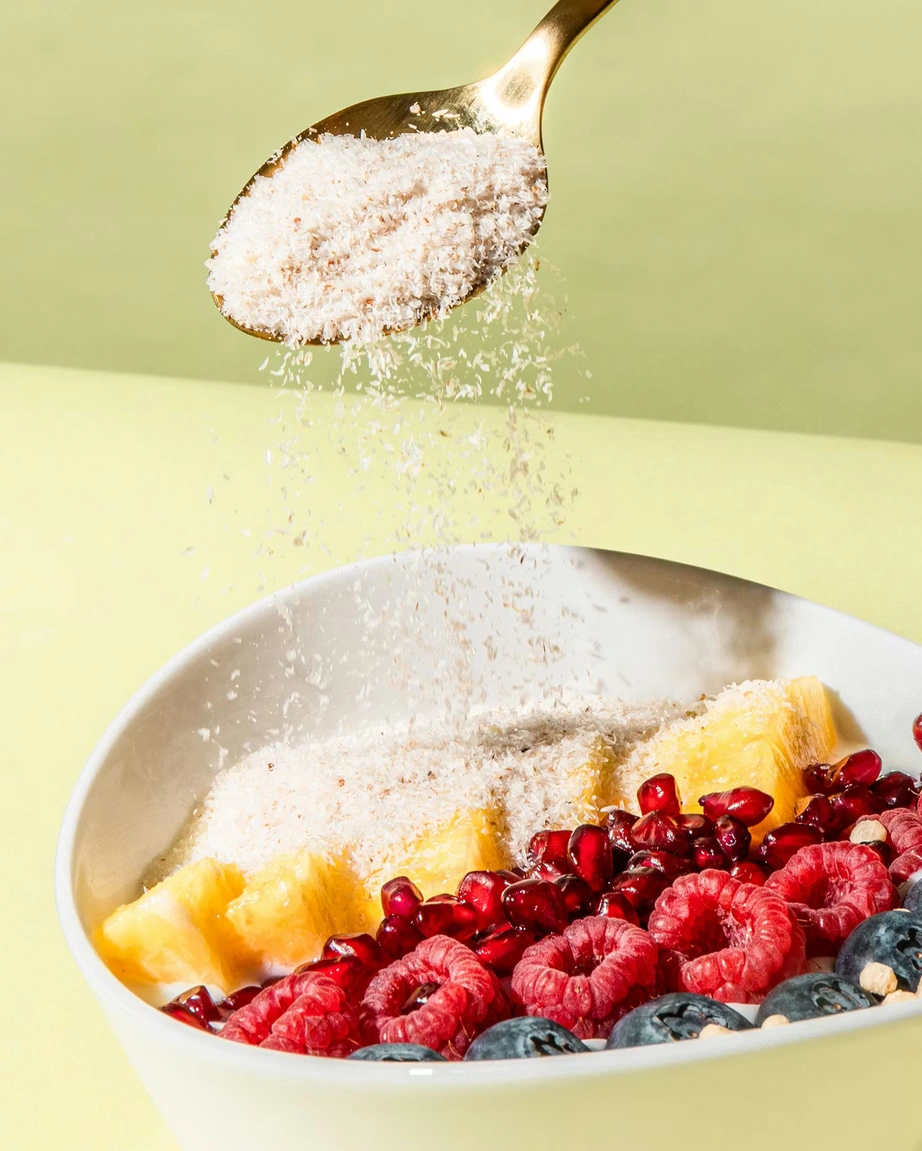 Ein goldener Löffel streut Flohsamenschalen über eine Frühstücksschale mit Ananas, Himbeeren, Heidelbeeren und Granatapfelkernen.