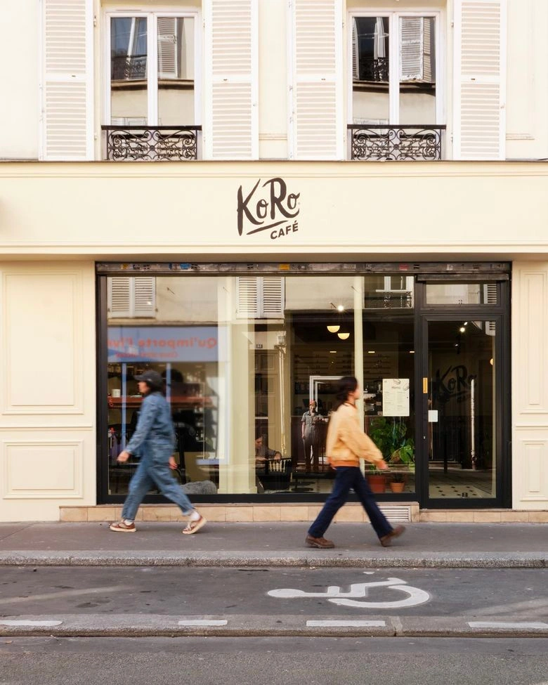 Zwei Passanten gehen an der großen Fensterfront des hellen „KoRo Café“ vorbei, das sich im Erdgeschoss eines klassischen Pariser Gebäudes befindet.