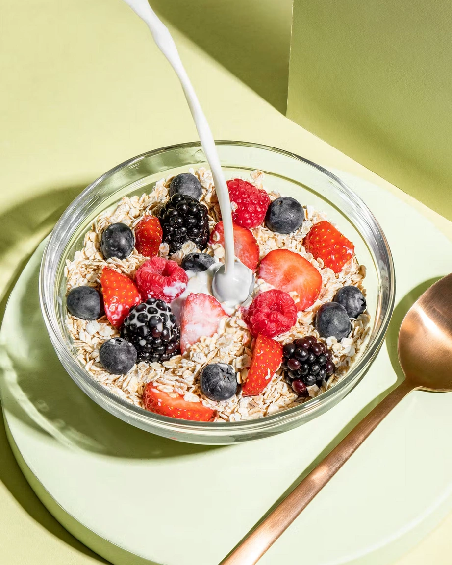 Nahaufnahme: Pflanzliche Milch wird über Bio-Haferflocken Kleinblatt und frische Beeren in einer Glas-Schale gegossen, daneben liegt ein kupferfarbener Löffel.