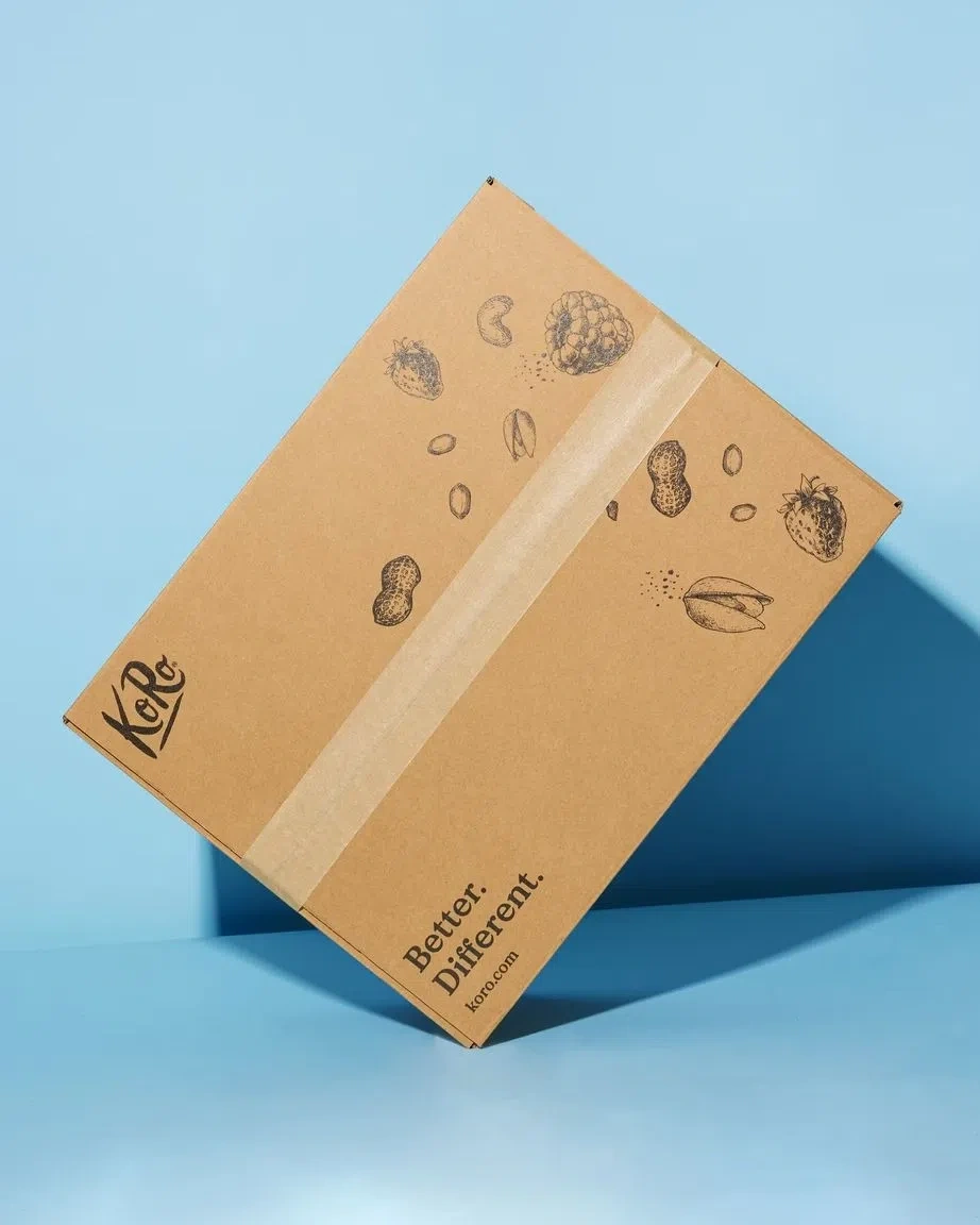 A brown shipping box, printed with the KoRo logo in black on the left side, various graphic depictions of nuts and fruits (pistachios, cashews, raspberries), and the text “Better. Different. koro.com” at the bottom. The box floats diagonally against a light blue background.
