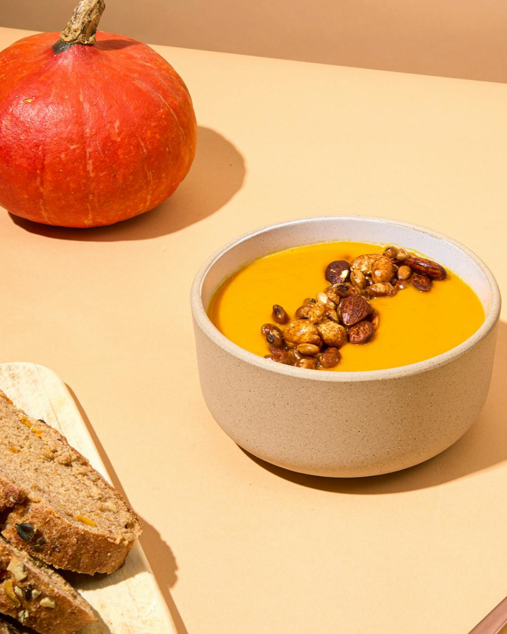 Eine dicke, leuchtend orangefarbene Kürbissuppe in einer beigefarbenen Keramikschüssel. Die Suppe ist mittig mit einer Mischung aus gerösteten Nüssen und Samen garniert. Im oberen linken Teil des Bildes liegt ein ganzer, oranger Hokkaido-Kürbis, und links unten sind Scheiben von Vollkornbrot auf einem Holzbrett angeschnitten.