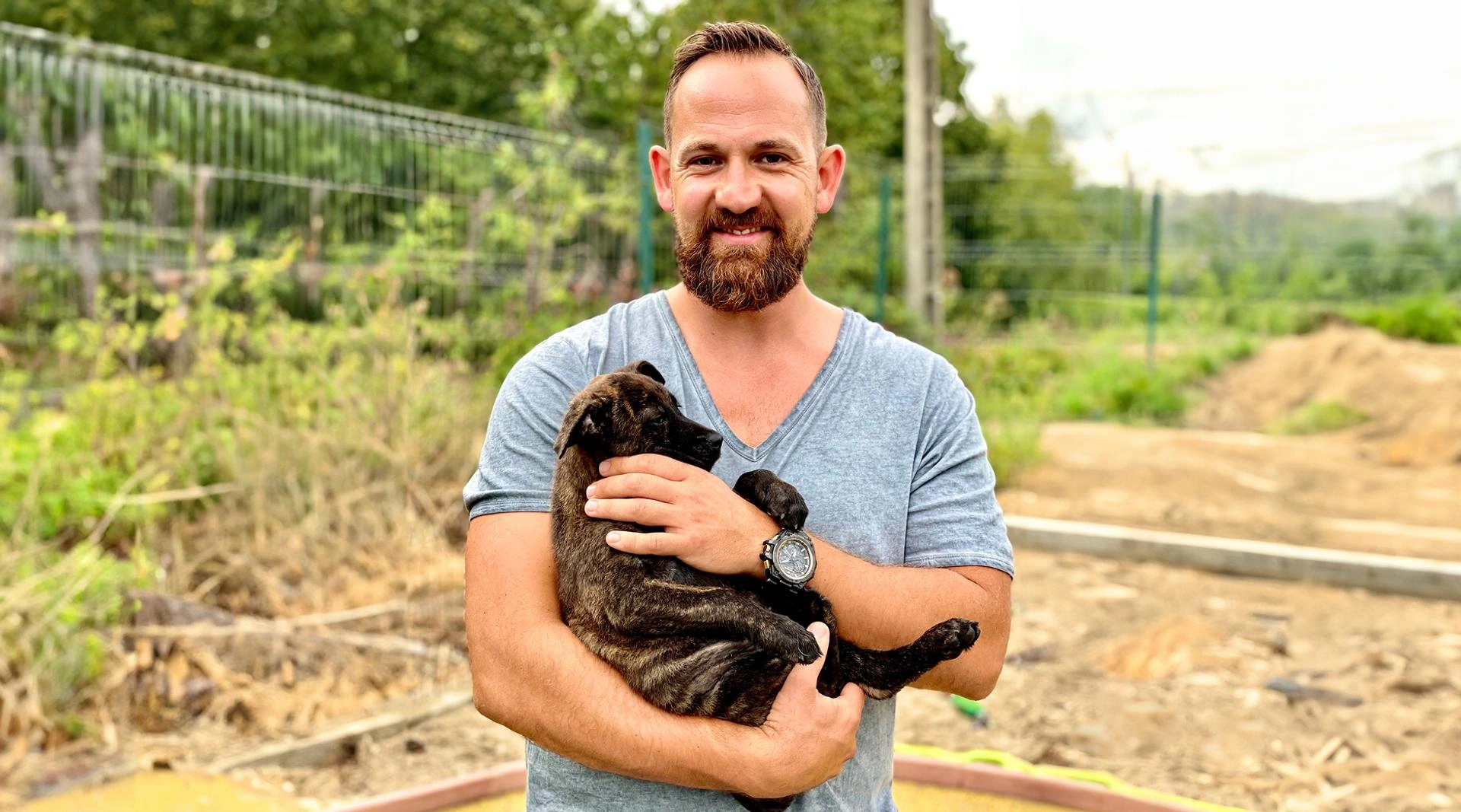 Mann mit kurzem Haar und Bart hält einen dunkel gestreiften Hundewelpen im Arm, Hintergrund mit Zaun und Grünfläche
