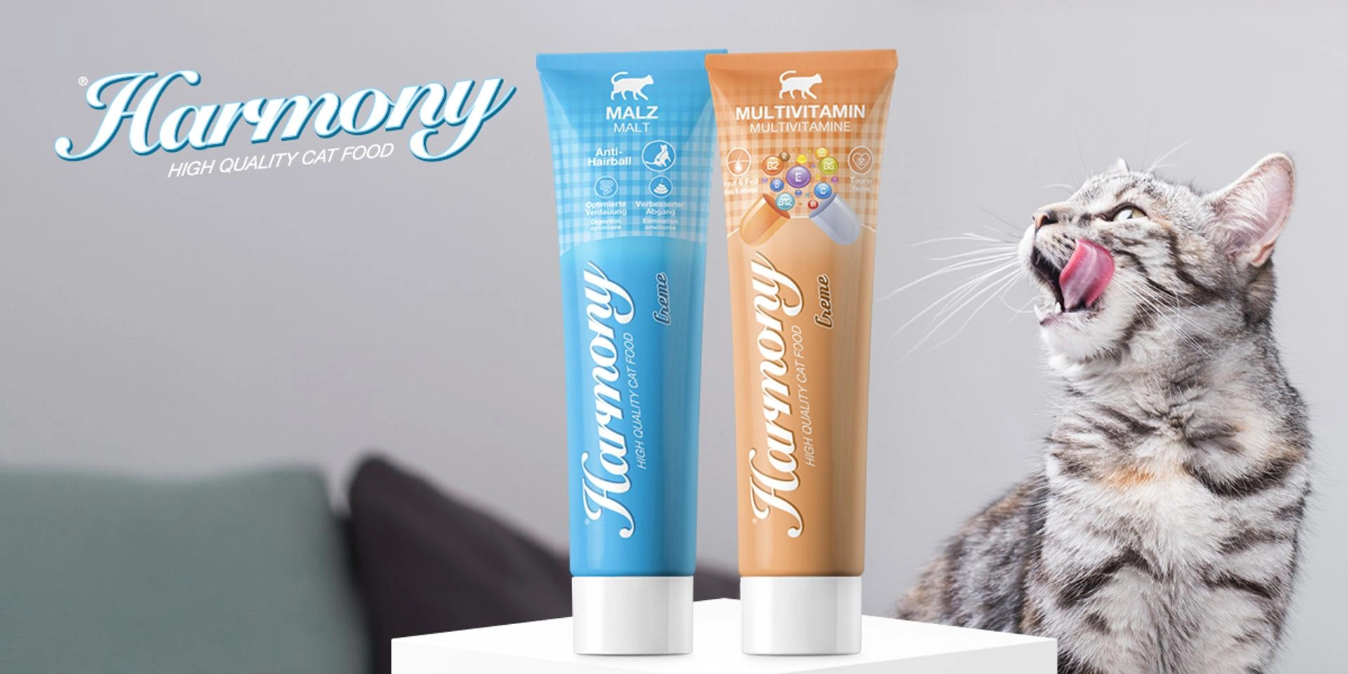 Zwei Harmony Paste Tuben Malz und Multivitamin auf weissem Podest, rechts graue Katze mit ausgestreckter Zunge, Harmony Logo oben links