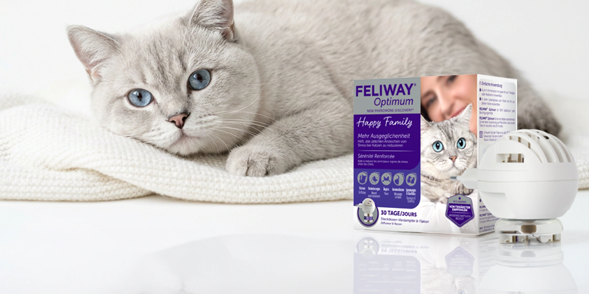 FELIWAY Optimum Happy Family Verdampfer und Nachfüllflakon neben einer entspannten Katze mit blauen Augen auf heller Decke in ruhiger Wohnatmosphäre.