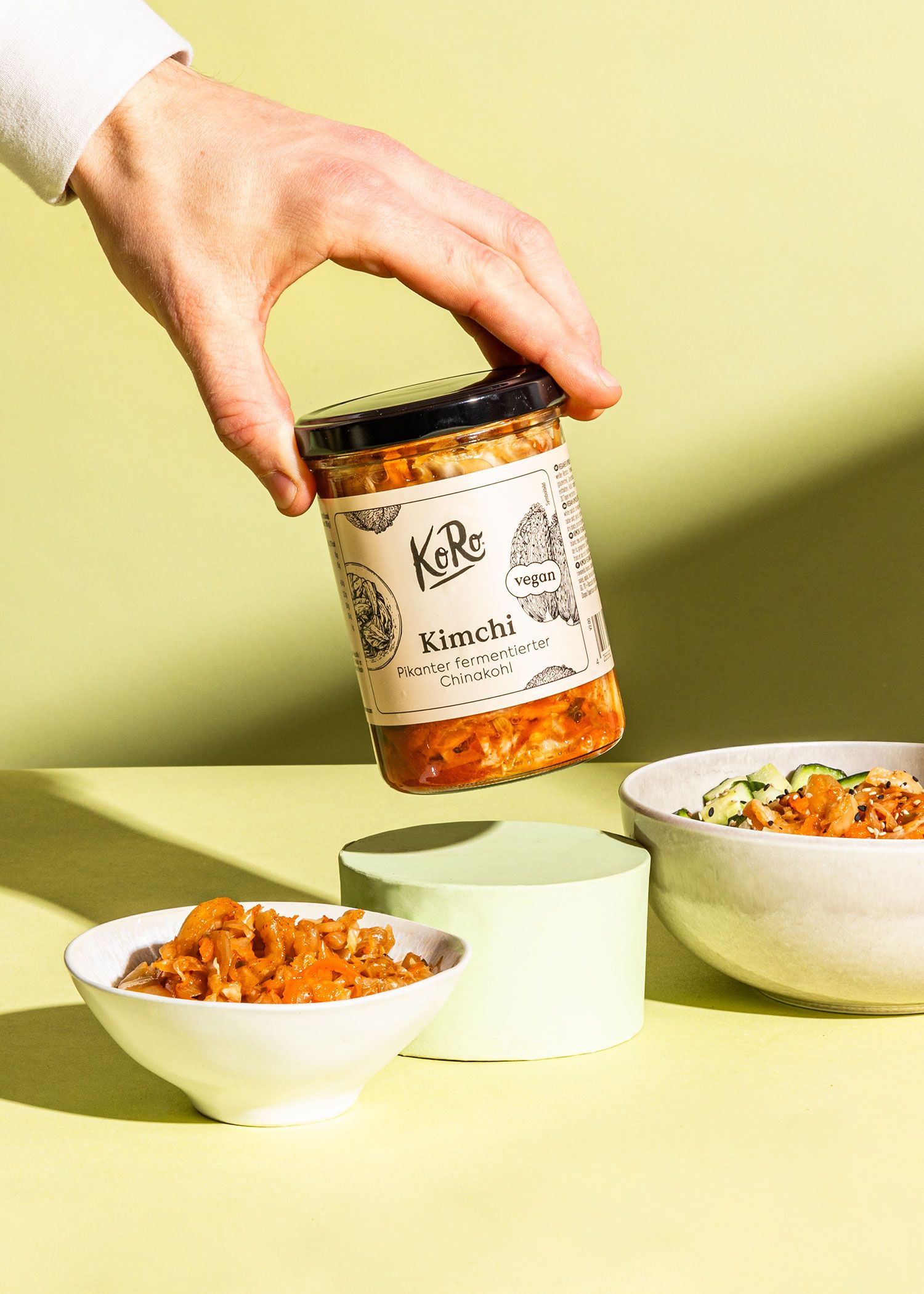 Hand hält ein Glas mit Kimchi, daneben zwei Schalen mit serviertem Kimchi