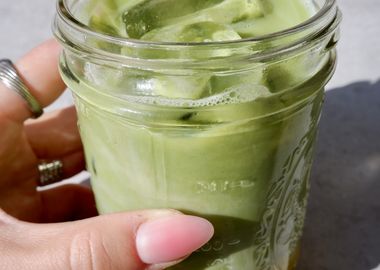 Hand hält ein Glas Iced Mango Matcha Latte mit Eiswürfeln; das Getränk zeigt eine grüne Matcha-Schicht über einer gelben Mangoschicht, im Sonnenlicht aufgenommen auf grauem Untergrund.