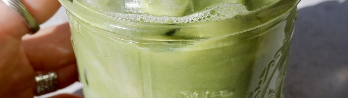 Hand hält ein Glas Iced Mango Matcha Latte mit Eiswürfeln; das Getränk zeigt eine grüne Matcha-Schicht über einer gelben Mangoschicht, im Sonnenlicht aufgenommen auf grauem Untergrund.