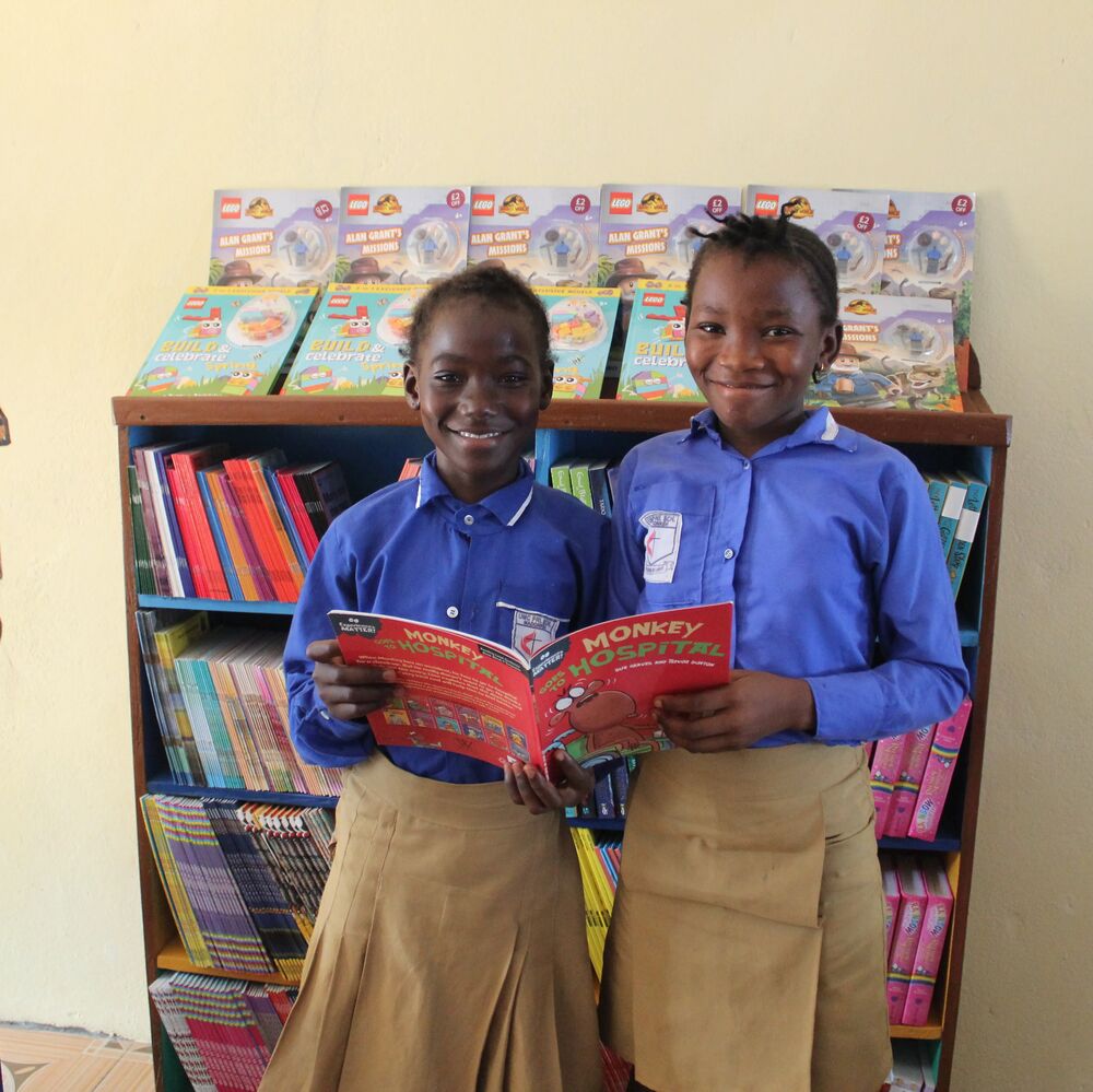 Smilende barn deler bok foran bokhylle i Explorer Library United Methodist Church Madonkeh i Sierra Leone