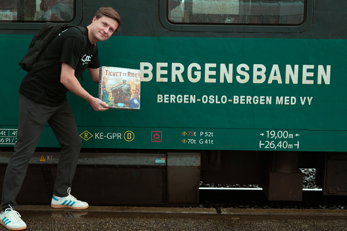 Outlands event-ansvarlig poserer med Ticket to Ride-spill foran Bergensbanen togvogn