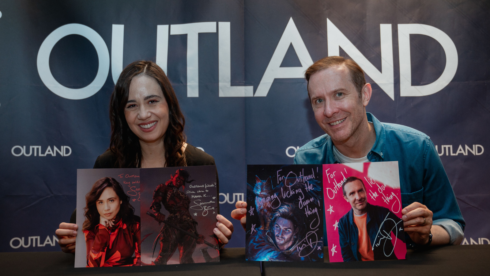 Samantha Béart og Tim Downie sitter foran Outland plakat med signerte bilder av seg selv og karakterene de spiller i Baldurs Gate 3 