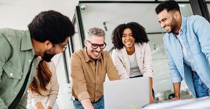 Five professionals gather around a silver laptop on a white table in a modern, well-lit office.