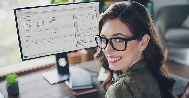A smiling woman with black-framed glasses sits at a wooden desk in a modern, well-lit office. 