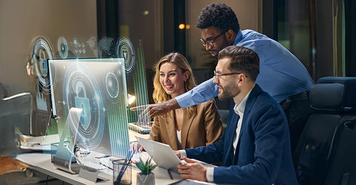 A group of three people collaborate in a modern office at night, working around a large transparent digital display showing futuristic security‑themed graphics, including a padlock icon and circular data visualizations.