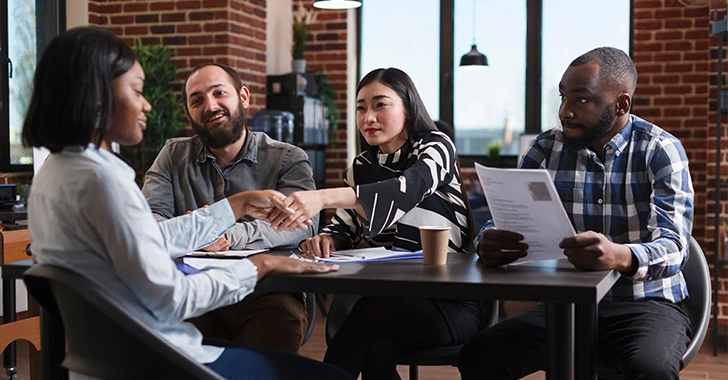 A diverse group of professionals seated around a table in a modern office, reviewing printed documents and exchanging papers during a collaborative meeting, with brick walls, large windows, and pendant lighting in the background.