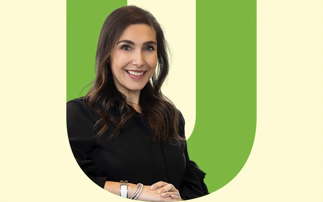 A smiling woman with long dark hair wearing a black shirt, posed against a light background with a green letter 'U'