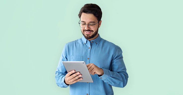 A man in a blue shirt smiles while using a tablet in front of a light green background. 