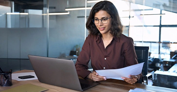A woman wearing glasses and a brown shirt sits at a wooden desk in a modern office.