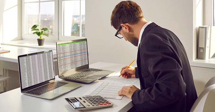 A man in a suit working at a desk with two laptops, a calculator, and paperwork. 