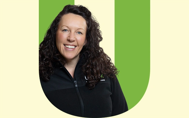 A woman with curly hair wearing a black zip-up jacket, smiling against a light green background. 