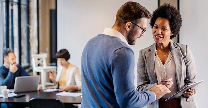 A man and woman discuss over a tablet in a bright office, with coworkers in the background