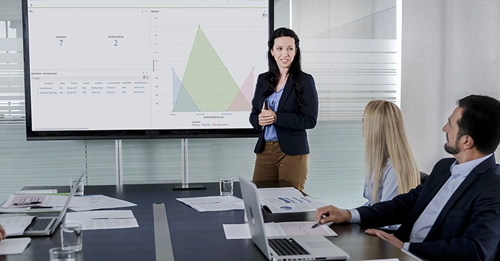 A woman presents data on a screen to a meeting room of people with laptops and papers. 
