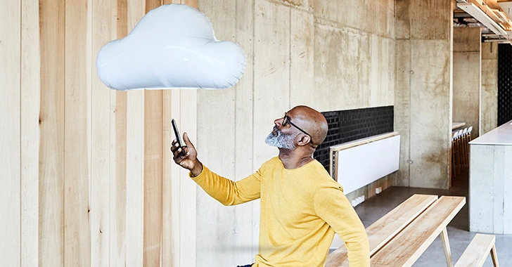 A man in a bright yellow sweater and glasses holds a smartphone while looking at a white cloud-shaped balloon in a modern indoor space with wooden and concrete elements.
