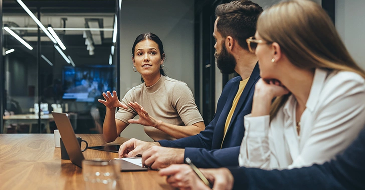 Three colleagues engaged in a business discussion around a polished wooden table in a modern office.