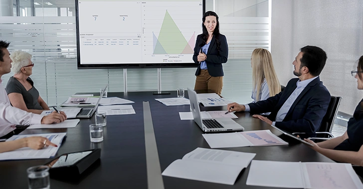 A group of professionals sit around a conference table during a meeting while a presenter stands at the front, gesturing toward a large screen displaying charts and graphs. 