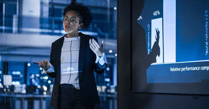 A professional presentation scene featuring a woman with dark curly hair and glasses, gesturing while explaining a bar chart projected on a screen.