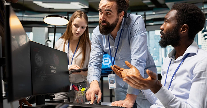 Three professionals in a modern office collaborate around a computer monitor displaying a 'CRITICAL ERROR' message. 
