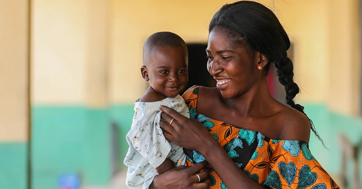 A smiling woman holds a baby, both with bright expressions, in a colorful setting. 