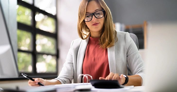 A professional woman with light brown hair and black-framed glasses works at a desk in a bright office.