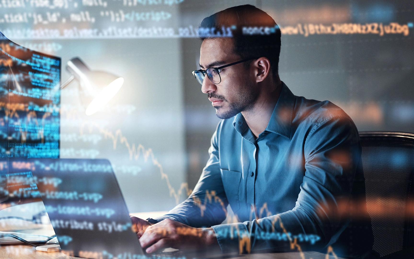 Person working intently on a computer in a dimly lit office, with reflections of code on the screen visible on the glass surface.