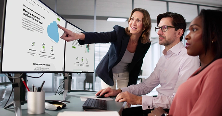 A professional office setting with three colleagues collaborating. One person stands, pointing at a monitor, while two others are seated, focused on the screens