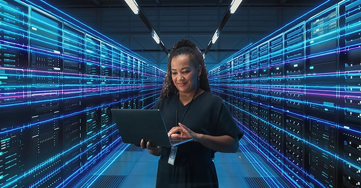 A woman in a black outfit using a laptop in a server room with glowing blue lights. 