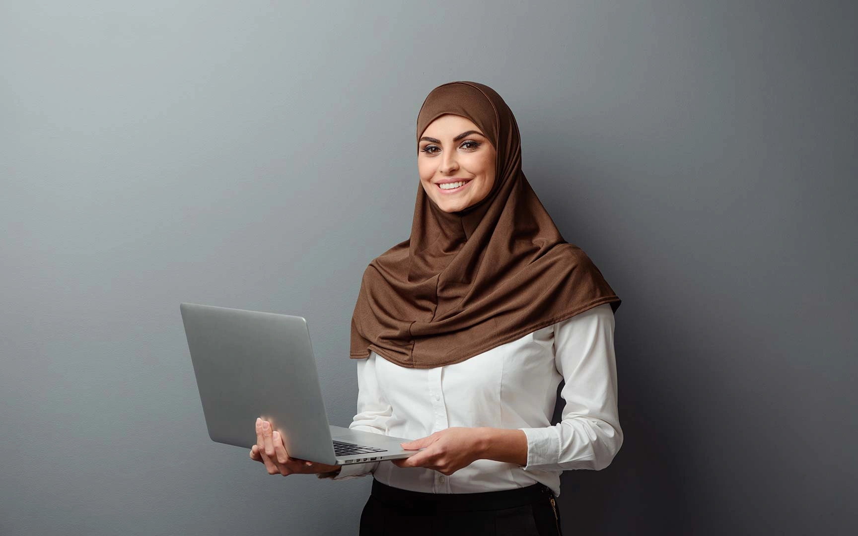 A woman wearing a brown hijab holds a laptop, smiling against a gray background. 