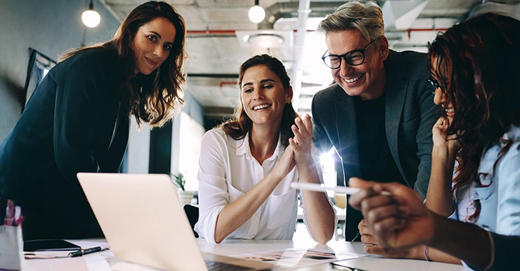 Four professionals smiling and engaging in a discussion around a laptop in a modern office setting.