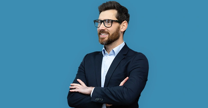 A man in a dark suit and glasses smiling, with arms crossed in front of a blue background. 