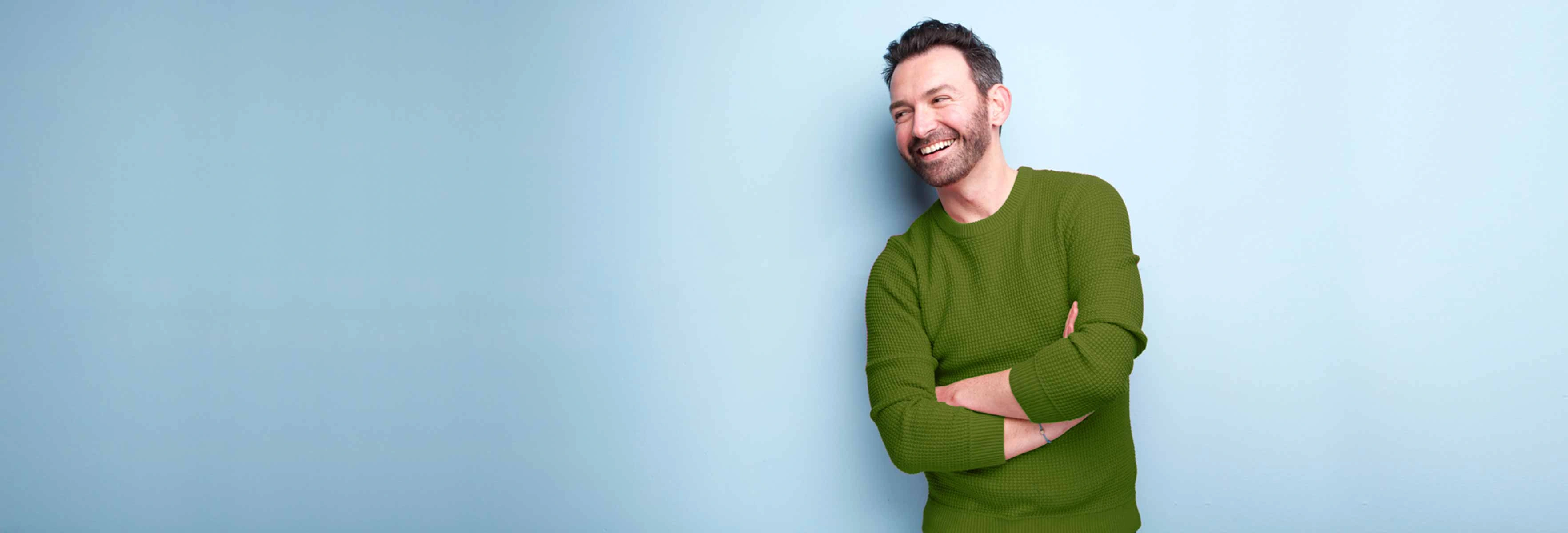 young man in green jumper with a smile on his face leaning against a blue background