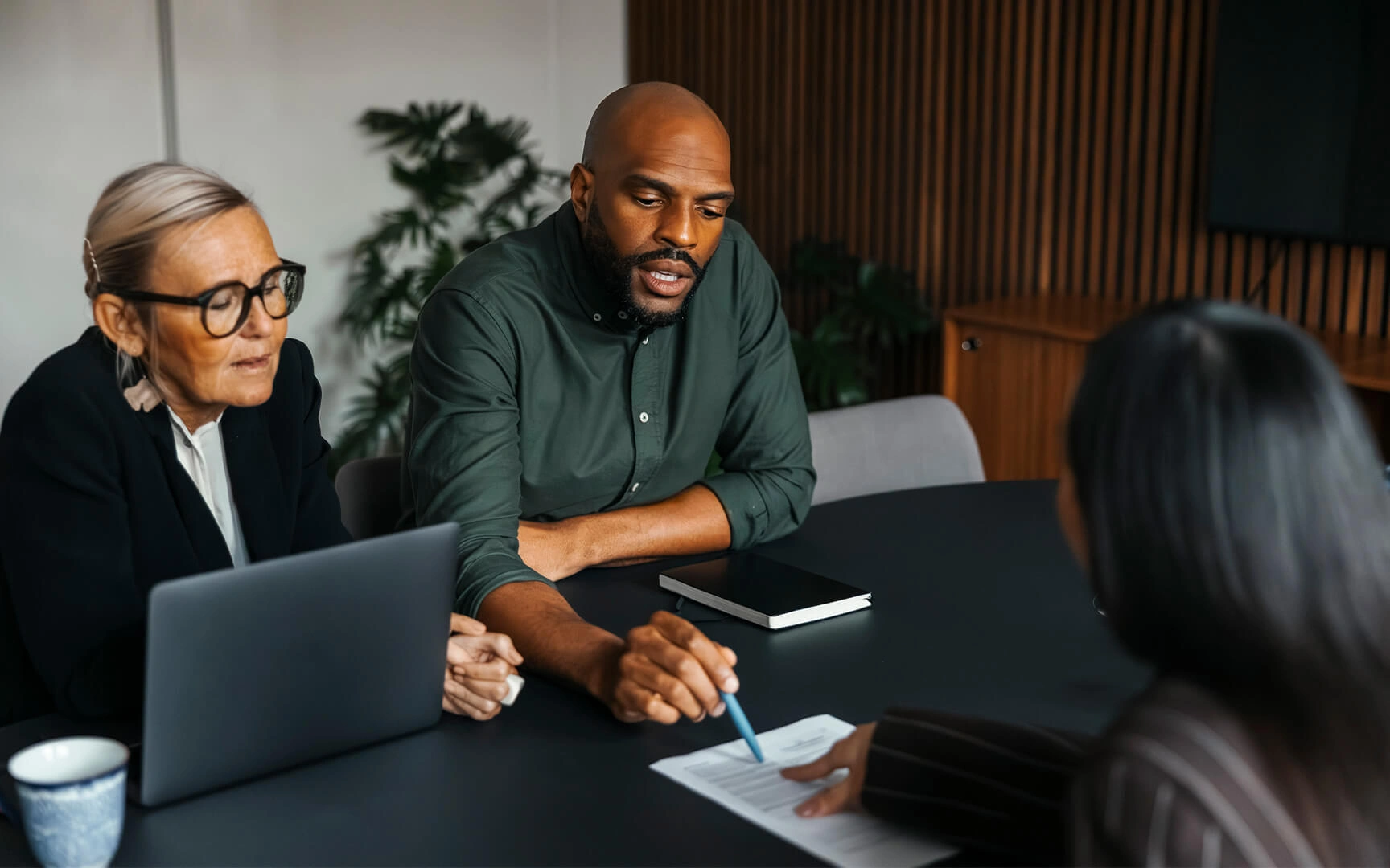 Three people seated at a meeting table reviewing a document together, with one person pointing at the page while another takes notes on a laptop.