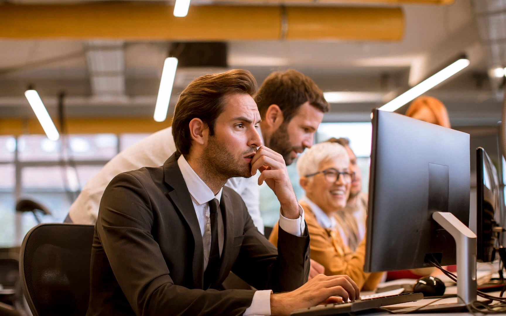 Professionelles Büroumfeld mit einer Gruppe von Mitarbeitenden an Desktop-Computern; eine Person im Vordergrund konzentriert sich auf einen Bildschirm, während Kollegen in einem modernen Arbeitsbereich mit warmer Beleuchtung zusammenarbeiten.