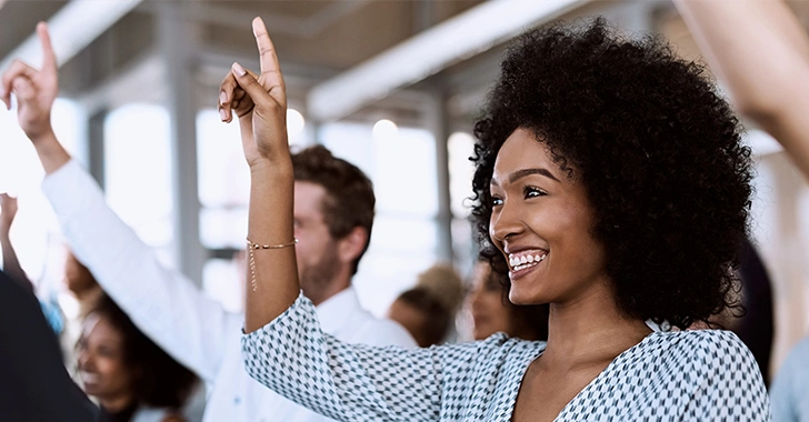 A smiling woman raises her hand enthusiastically during a business meeting or training session, with colleagues doing the same in the background.