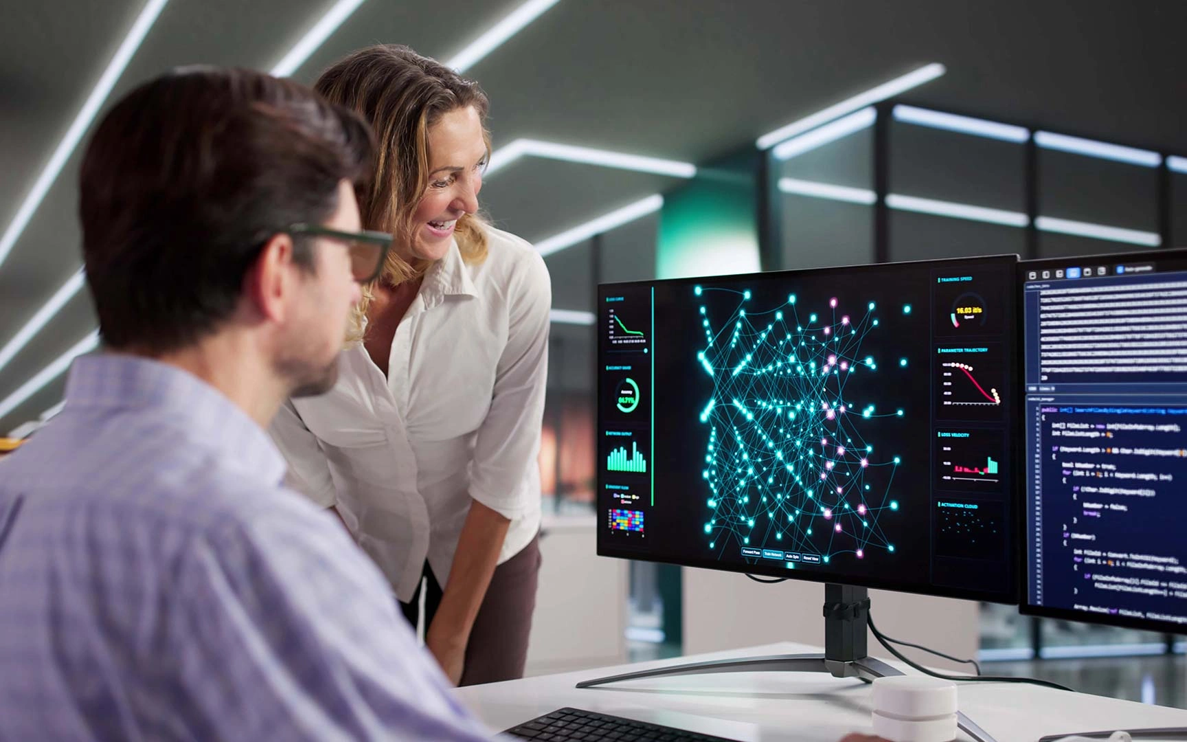 Deux professionnels collaborant à un bureau, examinant des visualisations de données et du code sur un écran large affichant un graphique de réseau et des tableaux de bord analytiques dans un bureau moderne.